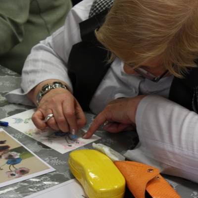 Zbliżenie na kobietę pochyloną nad białą kartką. W prawej dłoni kobieta trzyma pastel. Przed białąkartką kolorowy obrazek przedstwiający kwiaty w wazonie.