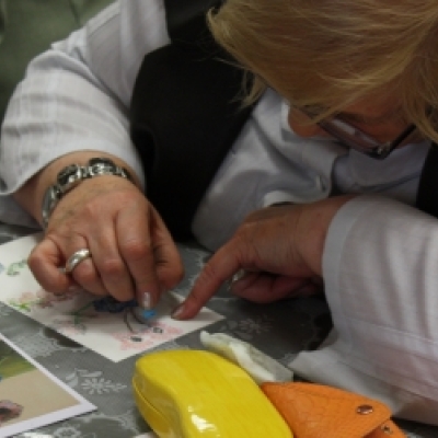 Kolorowe zdjęcie przedstawia kobietę siedzącą przy stole. Na stole leży reprodukcja obrazu. Kobieta koloruje ilustrację wzorując się na reprodukcji. 