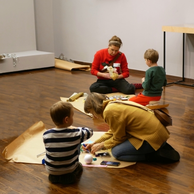  Cztery osoby znajdują się w jasnym pomieszczeniu na drewnianej podłodze. Na pierwszym planie kobieta w musztardowym swetrze klęczy i pracuje z materiałami plastycznymi, obok niej siedzi chłopiec w pasiastym swetrze. W tle kobieta w czerwonym swetrze siedzi po turecku, trzymając materiały, naprzeciwko niej chłopiec w zielonej bluzie i czerwonych spodniach siedzi tyłem. Wokół nich leżą arkusze papieru i różne materiały plastyczne.