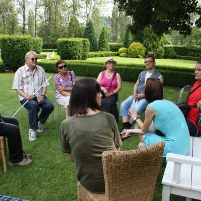 kilka osób dorosłych siedzi na krzesłach w kręgu wśród zielonych drzew